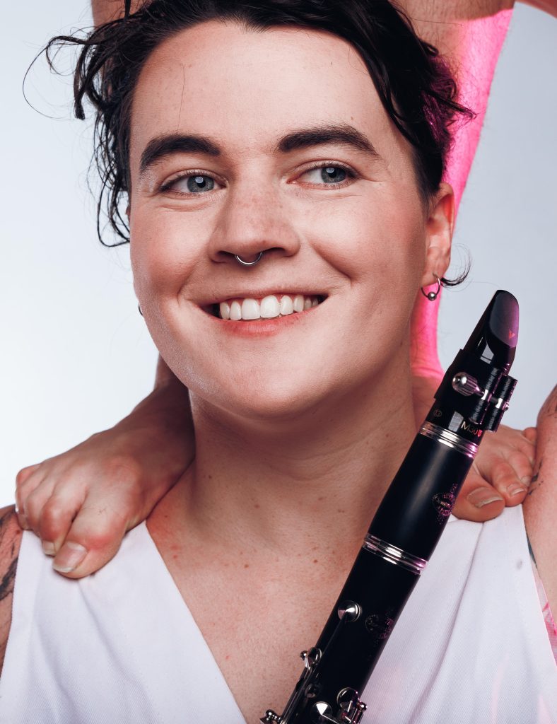 A close up portrait of Winter Chapman. She is a fair skinned woman with dark hair and blue eyes, wearing a white top. The feet of a person standing on her shoulders are visible. She is holding a clarinet, smiling and looking off to her left.
